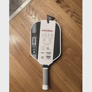 May include: A black and white pickleball paddle with a gray handle and a white grip. The paddle has a tag with the text "PERSEUS" and "JOOLA". The paddle is lying on a wooden surface.