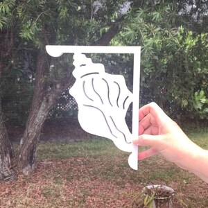 May include: White metal shelf bracket with a seashell design. The bracket is being held in a hand against a green leafy background.