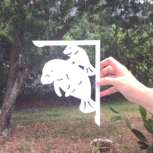 Op de afbeelding: Witte metalen wandbeugel met een uitgesneden ontwerp van twee lamantijnen. De lamantijnen kijken naar rechts en zijn op elkaar gelegd. De beugel is ontworpen om aan een muur te worden gemonteerd.