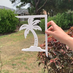 Op de afbeelding: Witte houten palmboom uitsnijding met een eenvoudig ontwerp. De uitsnijding is bevestigd aan een witte houten beugel. De beugel wordt vastgehouden door een hand.