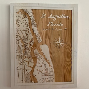 May include: A wooden map of St. Augustine, Florida, with a compass rose and the coordinates 29.9012° N, 81.3124° W. The map is framed in a white frame.