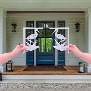 Op de afbeelding: Witte houten uitsnijding van twee vogels die in het water staan, vastgehouden voor een blauwe voordeur met glazen panelen. De deur bevindt zich aan een huis met witte bekleding en een veranda.
