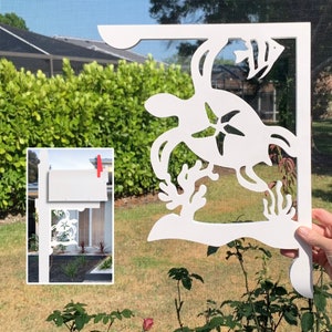 Op de afbeelding: Witte metalen brievenbusbeugel met een uitgesneden ontwerp van een zeeschildpad, een vis en koraal. De beugel is gemonteerd op een witte brievenbuspaal.