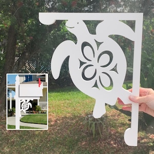 Op de afbeelding: Witte houten brievenbusbeugel in de vorm van een zeeschildpad met een bloemmotief. De beugel is ontworpen om een brievenbus op een paal te houden.