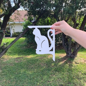 Op de afbeelding: Wit metalen kattenvormig wandrek. Het rek is gevormd als een zittende kat met zijn staart om zijn lichaam gekruld. Het rek is ontworpen om aan de muur te worden gemonteerd.