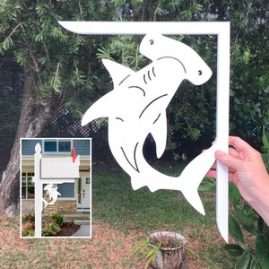 May include: White wooden mailbox post with a hammerhead shark design. The post is attached to a white wooden mailbox with a white flag.