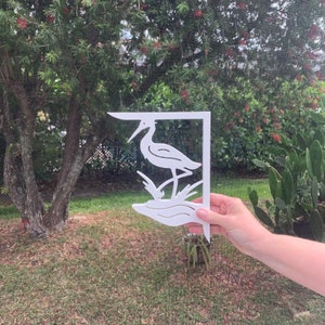 Op de afbeelding: Witte metalen tuinbeugel met een uitgesneden ontwerp van een reiger die in het water staat. De beugel wordt voor een groene gazon en een boom gehouden.