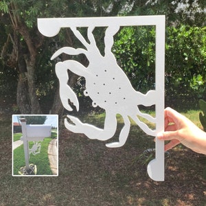 Op de afbeelding: Witte metalen brievenbusbeugel in de vorm van een krab. De beugel is bevestigd aan een witte brievenbuspaal. De beugel wordt getoond in de hand van een persoon die hem omhoog houdt.