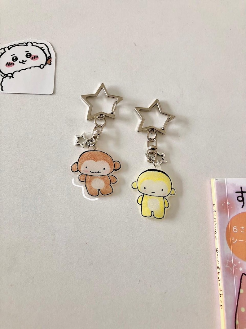 May include: Two keychains featuring star-shaped silver metal clasps and small monkey figures. One monkey is light brown with a white belly, and the other is yellow with a white belly. Both figures have simple facial features and rounded ears.