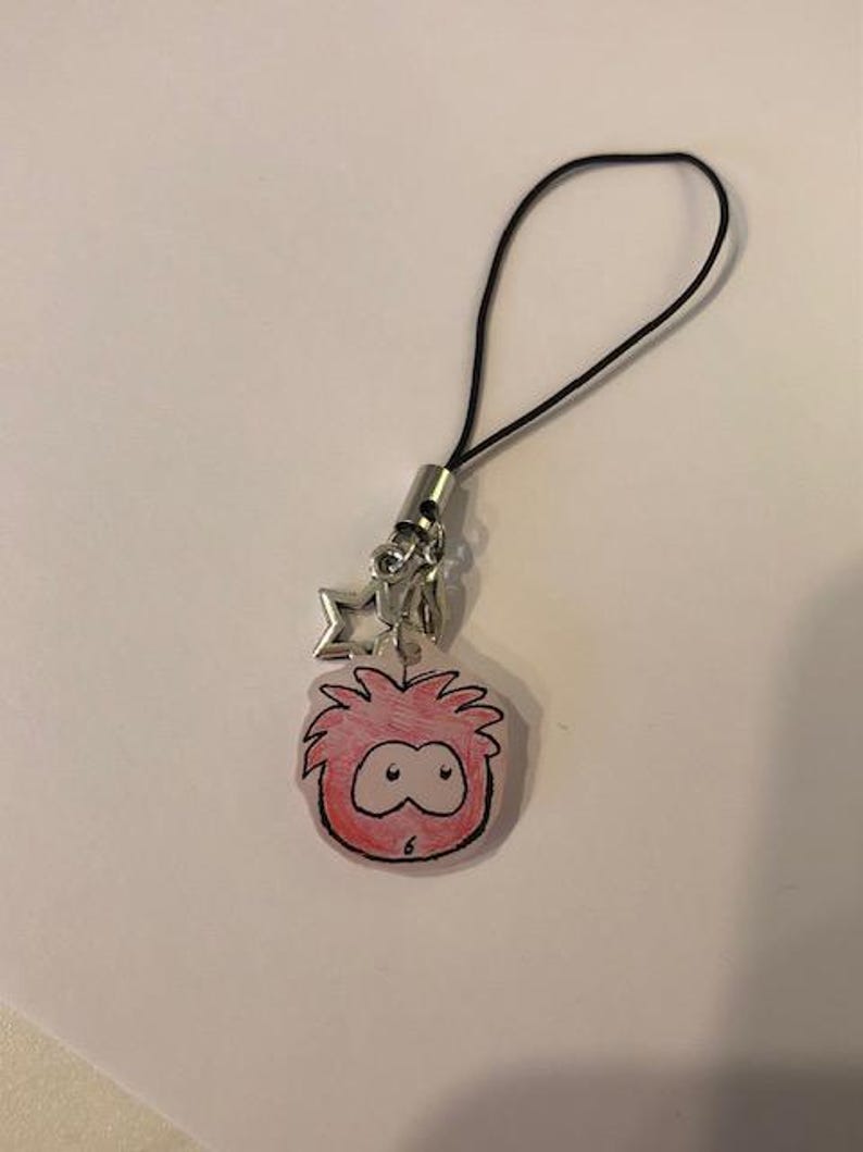 May include: A red and black cartoon character charm with a black cord and silver-colored metal accents. The charm features a star-shaped accent. The cartoon character has a surprised expression.