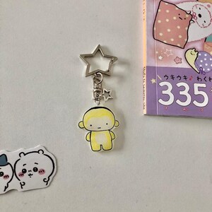 May include: A yellow and white cartoon character keyring featuring a star-shaped metal clasp. The character has a simple face with black eyes and a small mouth, attached to a silver-toned metal ring and a smaller star charm.