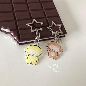 May include: Two keychains featuring silver star-shaped clasps attached to small, illustrated monkey charms. One monkey is yellow with a light belly, and the other is brown with a light belly. Both charms hang from a dark brown chocolate bar notebook.