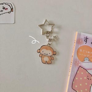 May include: A silver star-shaped keyring featuring a small, light brown monkey charm with a simple facial expression. The monkey has a round head, small ears, and a lighter-coloured belly. The keyring includes a secondary, smaller star charm.