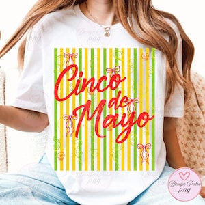 Pode incluir: Camiseta branca com um design de Cinco de Mayo. O design apresenta texto vermelho e laços decorativos sobre um fundo de listras verticais amarelas e verdes. Pequenos gráficos de caveiras também estão presentes.