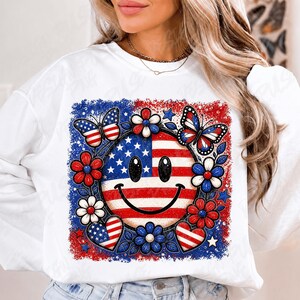 May include: White sweatshirt featuring a patriotic design. The graphic includes a smiley face with the American flag, surrounded by red, white, and blue flowers, butterflies, and hearts. The design has a distressed, vintage look.
