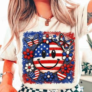 May include: White t-shirt featuring a patriotic smiley face design. The design includes a smiley face with the American flag, surrounded by red, white, and blue flowers, butterflies, and hearts. The design is a festive and colorful representation of the USA.