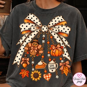 May include: A charcoal gray t-shirt featuring a large graphic of a white bow with black polka dots. Hanging from the bow are charms including a turkey, a pumpkin, a slice of pie, a mug labeled GIVE THANKS, a sunflower, an acorn, and leaves.