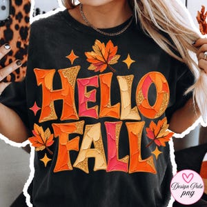 May include: A black t-shirt featuring the text HELLO FALL in a textured, impasto-style font with shades of orange, red, and cream. The design includes glittery gold accents, stylised autumn leaves, and four-pointed star shapes surrounding the text.