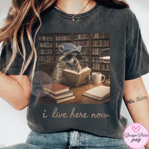 May include: Dark gray t-shirt featuring a raccoon wearing sunglasses and a cap, reading a book in a library setting. The text "i live here now" is printed below the image. A mug and books are on the table.