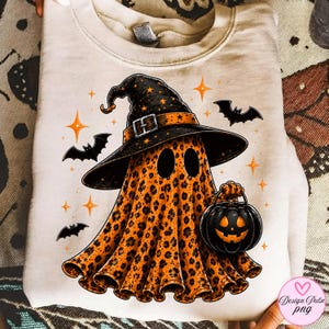 May include: A beige crewneck sweatshirt featuring a graphic of a ghost wearing a black witch hat with a buckle. The ghost has an orange leopard print pattern and holds a black jack-o'-lantern. The design includes small black bats and orange stars.