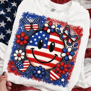 May include: White sweatshirt featuring a patriotic smiley face design. The design incorporates the American flag, butterflies, flowers, and hearts in red, white, and blue. The smiley face has a happy expression.