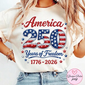 Puede incluir: Camiseta color crema con un dise&ntilde;o patri&oacute;tico que celebra el 250 aniversario de Am&eacute;rica. El dise&ntilde;o presenta la palabra "America" en rojo, el n&uacute;mero "250" con un patr&oacute;n de estrellas y rayas, y el texto "Years of Freedom 1776-2026" en azul.