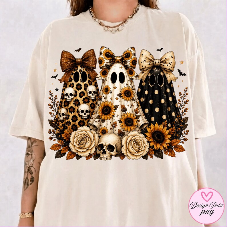 May include: A cream-colored t-shirt featuring a graphic of three ghosts wearing large bows. The ghosts are decorated with leopard print, sunflowers, and gold polka dots. The design includes small skulls, sunflowers, roses, and bats.