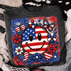 May include: A dark gray sweatshirt featuring a patriotic design. The design includes a smiley face with the American flag, surrounded by red, white, and blue flowers, butterflies, and hearts. The sweatshirt is a casual style.