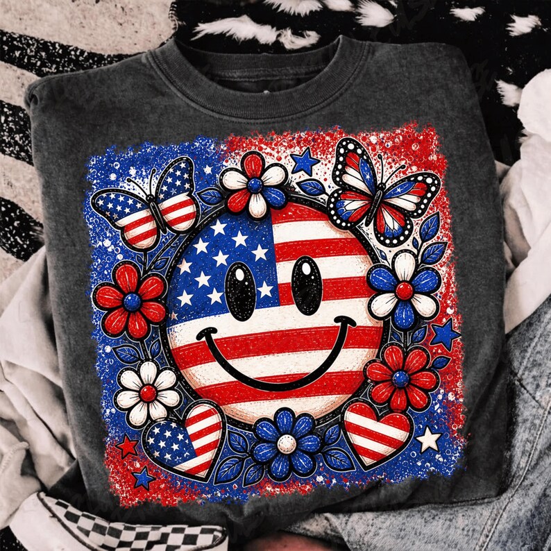 May include: A dark gray sweatshirt featuring a patriotic design. The design includes a smiley face with the American flag, surrounded by red, white, and blue flowers, butterflies, and hearts. The sweatshirt is a casual style.