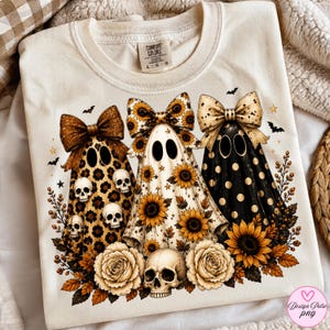 May include: A cream-colored t-shirt featuring a graphic of three ghosts wearing large bows. The first ghost has a leopard print, the second has a sunflower pattern, and the third has black and gold polka dots. Skulls, sunflowers, and roses surround them.