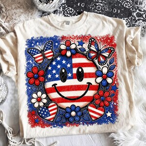 May include: Cream-colored t-shirt featuring a patriotic smiley face design. The design includes the American flag, butterflies, flowers, and hearts in red, white, and blue. The shirt has a crew neck and short sleeves.
