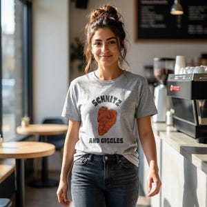 May include: A gray t-shirt with the words "Schnitz" above a fried chicken graphic and the words "And Giggles" below. The shirt is worn by a person in a cafe setting, paired with dark wash jeans.