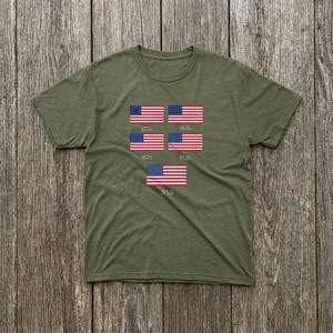 May include: Olive green t-shirt featuring a series of American flags from different historical periods. The flags are displayed with the years 1776, 1826, 1876, 1926, and 2020 below each flag. The shirt is made of a soft material.