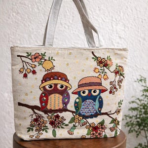 May include: Beige tote bag featuring an embroidered design of two owls wearing hats, perched on a branch with flowers. The owls have colorful details, and the bag has light gray handles. The bag is sitting on a wooden stool.