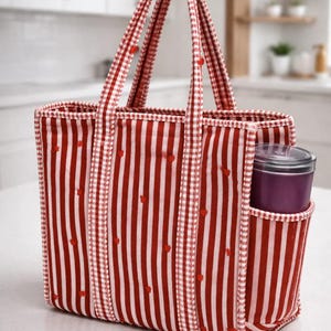 May include: Red and white striped tote bag with a gingham trim and handles. The bag has a side pocket holding a purple container. Small red heart-shaped accents are scattered across the stripes. The bag is designed for carrying items.