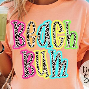 May include: A peach-coloured t-shirt featuring the words "Beach Bum" in a leopard print design with neon outlines. The shirt is a casual style, suitable for a day at the beach or a summer outing.