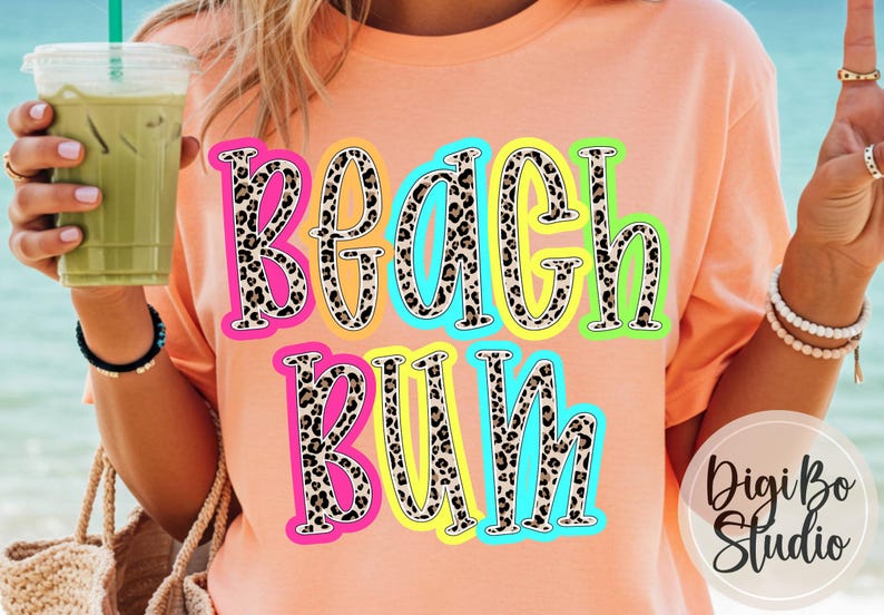 May include: A peach-coloured t-shirt featuring the words "Beach Bum" in a leopard print design with neon outlines. The shirt is a casual style, suitable for a day at the beach or a summer outing.