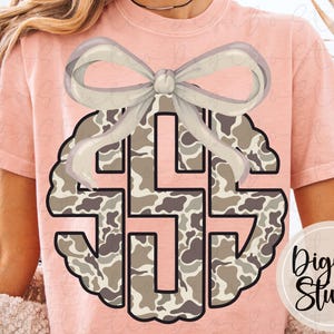 May include: A light pink t-shirt featuring a monogram design with a camouflage pattern in shades of brown and gray. A light gray bow is placed above the monogram. The text "DigiBo Studio" is in a circle on the bottom right.