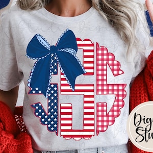 May include: A light grey t-shirt with a patriotic design. The design features a large blue bow with scalloped edges, and the letters "USA" filled with red and white stripes and stars. The design is in a circular shape.