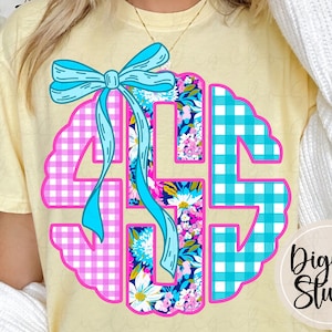 May include: A light yellow t-shirt featuring a monogram design. The monogram is a scalloped circle with the letters GBS in a pink gingham pattern, blue and white gingham, and floral print. A blue bow and ribbons are at the top.