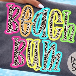May include: A dark grey t-shirt with the words "Beach Bum" in a colourful, playful design. The text features a leopard print pattern with bright pink, yellow, and blue outlines. The shirt is held by a person with a blue background.