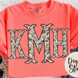 May include: Coral-colored t-shirt with the letters K, M, and H in a camouflage pattern. The letters have a decorative, ornate design. The shirt also features the text "DigiBo Studio" in a circular design.