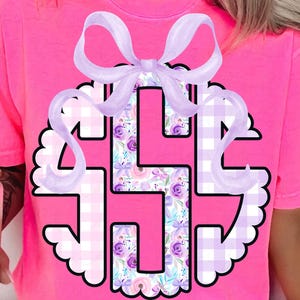 May include: A vibrant pink t-shirt with a large monogram design. The monogram incorporates floral and gingham patterns. A light purple bow adorns the top of the monogram, creating a decorative element.