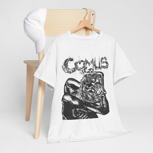 May include: A white short-sleeve t-shirt featuring a black and white graphic print of a hunched, dark figure with the text COMUS in a distressed font above it, accompanied by a plain white baseball cap hanging on a wooden hanger.