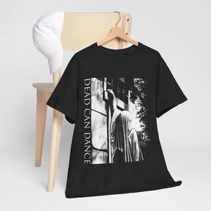 Könnte beinhalten: Ein schwarzes Kurzarm-T-Shirt mit einem weißen Grafikdruck einer drapierten Statue vor einer Steinmauer. Der Text "DEAD CAN DANCE" ist vertikal in weißen Großbuchstaben auf der linken Seite des rechteckigen Grafikdesigns aufgedruckt.