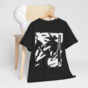 May include: A black short-sleeve t-shirt featuring a high-contrast white graphic print of a face and the text CLOCK DVA in a vertical rectangular border. A plain white baseball cap is also present, hanging on the back of a wooden chair.