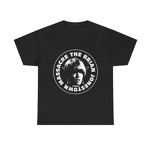 Può includere: T-shirt nera con grafica circolare bianca. La grafica presenta il testo "MASSACRE THE BRIAN JONESTOWN" attorno a un ritratto. Sono inclusi anche una stella e un ritratto di una persona.