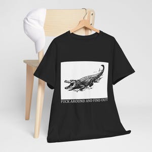 Könnte beinhalten: Ein schwarzes Kurzarm-T-Shirt mit einer weißen rechteckigen Grafik, die eine schwarz-weiße Illustration eines Alligators zeigt. Unter dem Alligator steht der Text FUCK AROUND AND FIND OUT in einer schwarzen Serifenschrift. Eine weiße Baseballkappe ist dabei.