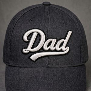 Könnte beinhalten: Dunkelgraue Baseballkappe mit dem Wort "Dad" in weißer Schrift auf der Vorderseite gestickt. Die Kappe hat einen gebogenen Schirm und einen Knopf oben. Ein stilvolles Accessoire für jeden Vater.