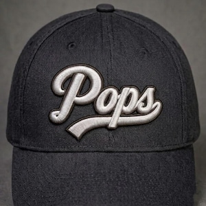 May include: A dark charcoal gray baseball cap featuring the word Pops embroidered in a white script font with a dark outline across the front panels. The cap has a structured crown, a curved brim, and visible stitching details throughout the fabric.